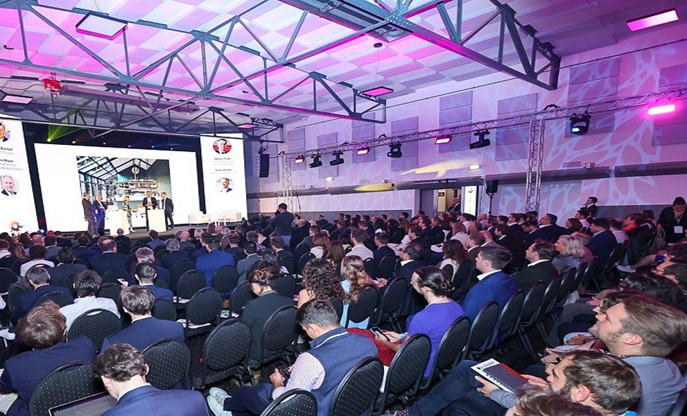 Eine Eventhalle voller Publikum, in der Ferne eine Bühne mit drei Personen und LED-Walls im Hintergrund