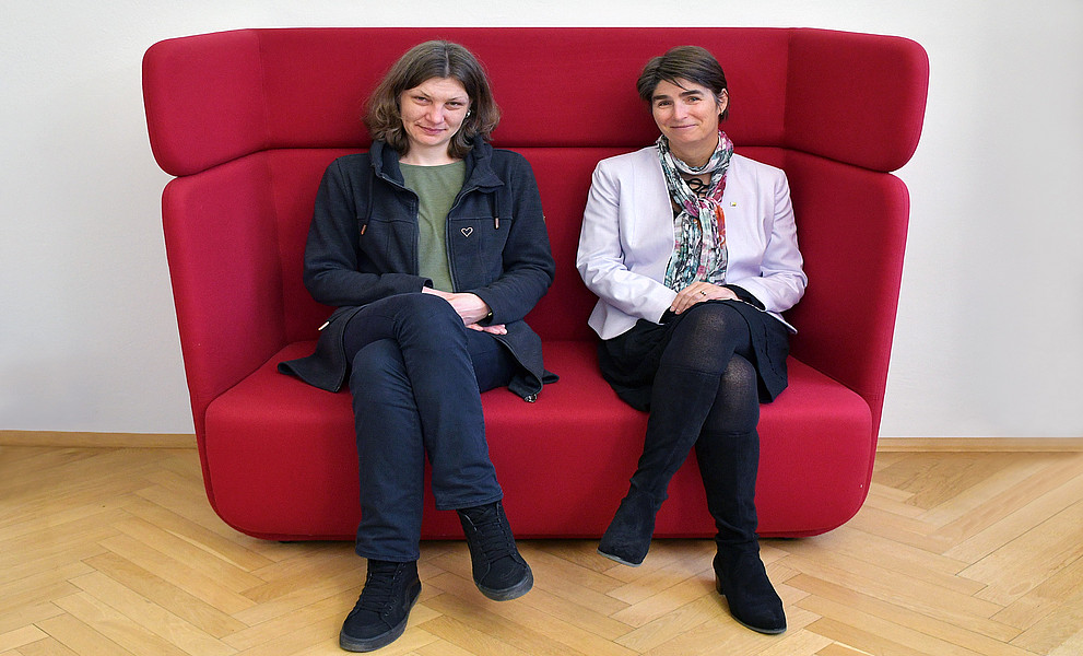 Sarah Zapusek und Mireille van Poppel nebeneinander sitzend auf einer roten Couch mit hoher Rückenlehne