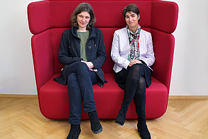 Sarah Zapusek and Mireille van Poppel sitting next to each other on a red couch with high backrests