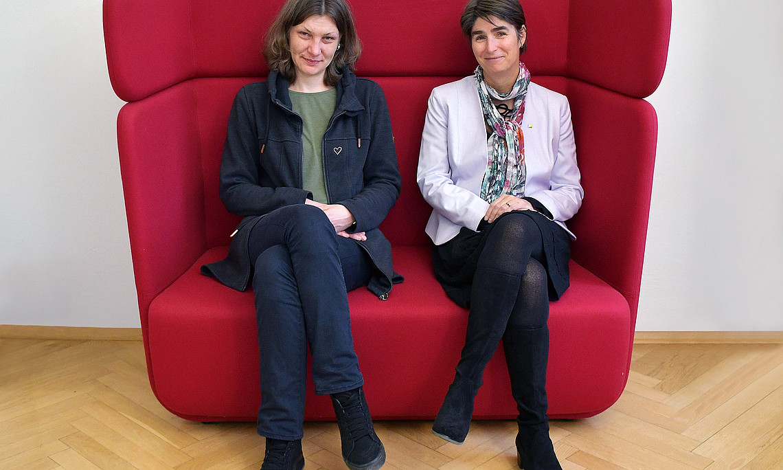Sarah Zapusek and Mireille van Poppel sitting next to each other on a red couch with high backrests