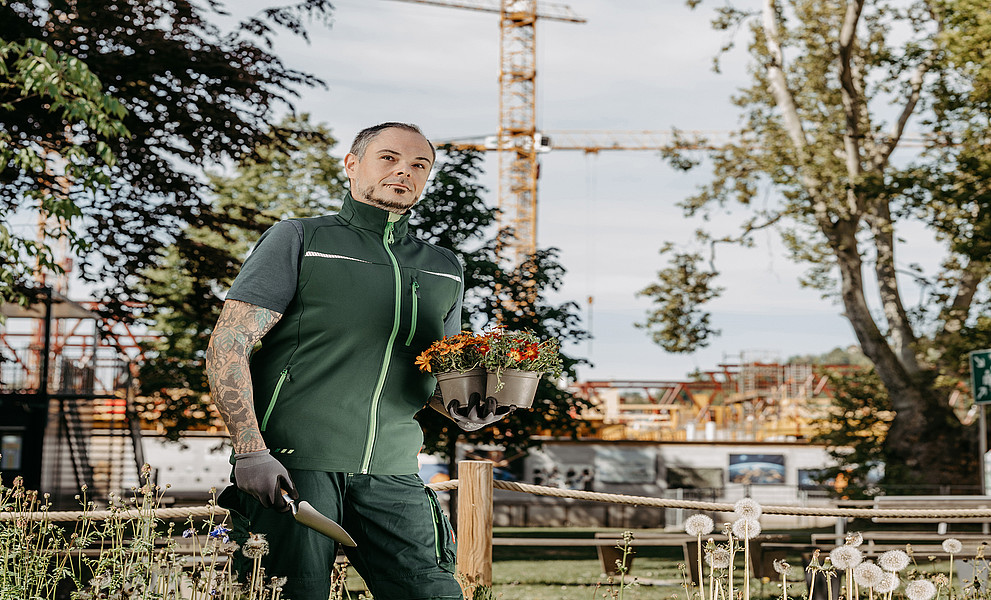 Gärtner Thomas Galik sitzt mit Schaufel und Pflanze in der Hand auf einem bepflanzten Trog