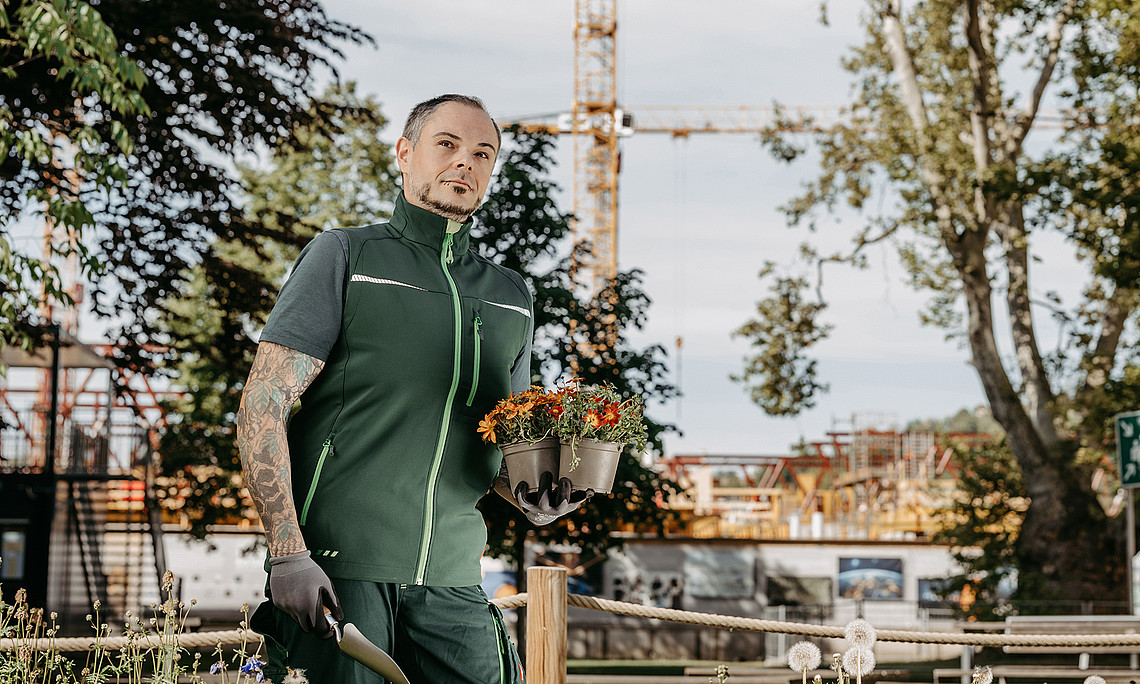 Gärtner Thomas Galik sitzt mit Schaufel und Pflanze in der Hand auf einem bepflanzten Trog
