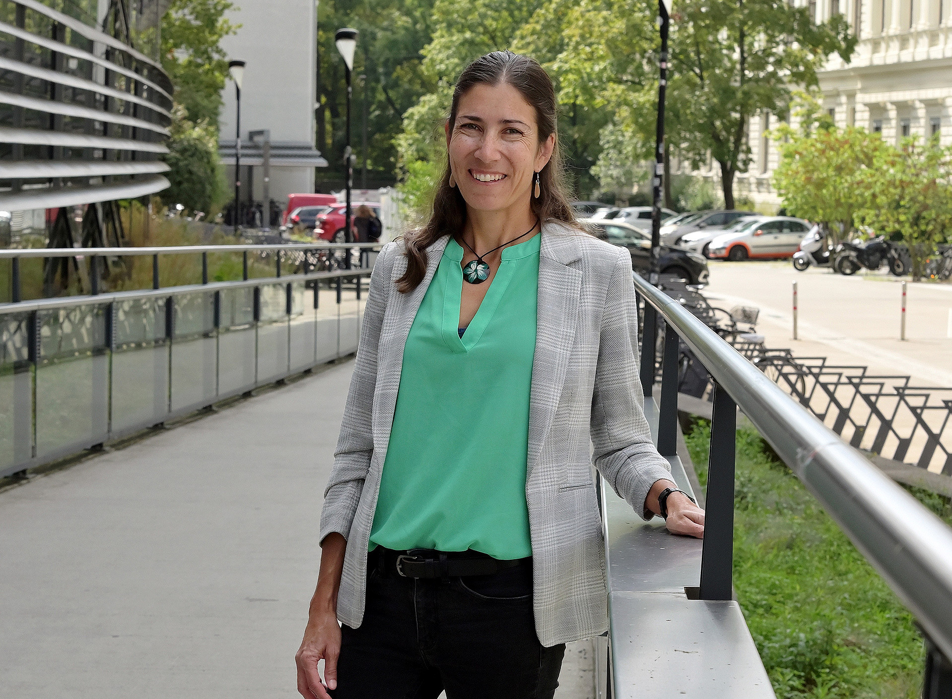 A middle-aged woman wearing a green top and a grey blazer is standing by a railing on a pavement on the University of Graz campus, surrounded by trees and parked cars ©University of Graz / Pichler