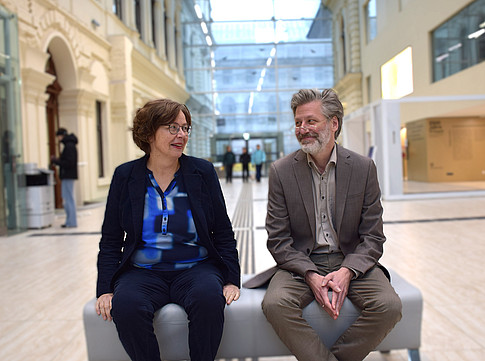 Edeltraud Aspöck and Julian Blunk sit on a bench in the foyer of the University Library of Graz and look at each other. 