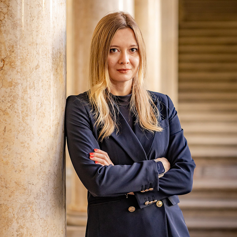 Das Foto zeigt eine Person mit langen, blonden Haaren und einem nachdenklichen Ausdruck. Sie trägt ein dunkelblaues, stilvolles Jackett und hat die Arme verschränkt. Die Person steht neben einer Marmorsäule, was auf eine Umgebung mit klassischen architektonischen Elementen schließen lässt. Das weiche Licht und die warmen Farbtöne verleihen dem Bild eine einladende Atmosphäre. 