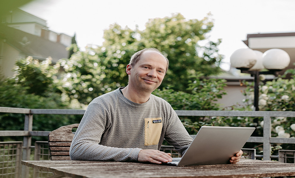 Tobias Stern sitzt vor seinem Laptop auf der Terrasse seines Instituts, im Hintergrund sind Büsche.
