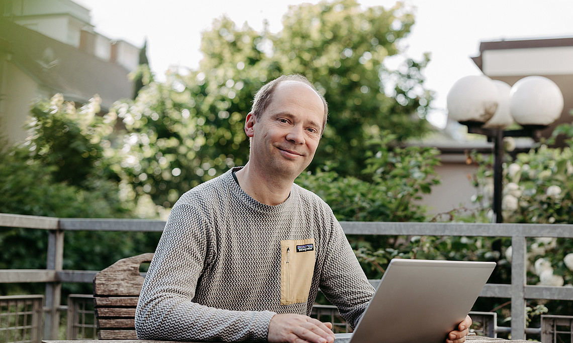 Tobias Stern sitzt vor seinem Laptop auf der Terrasse seines Instituts, im Hintergrund sind Büsche.