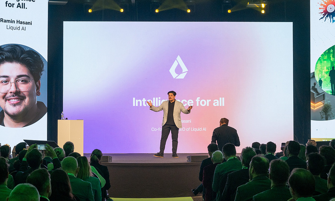 A speaker stands on a stage in front of a large screen displaying the words “Intelligence for all” and a logo. The background of the screen is designed in a color gradient from purple to pink. The audience sits in rows and listens to the presentation. Vertical banners with graphics and text are attached to the left and right of the stage. The stage lighting is bright, while the audience area is darker.