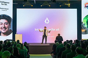 A speaker stands on a stage in front of a large screen displaying the words “Intelligence for all” and a logo. The background of the screen is designed in a color gradient from purple to pink. The audience sits in rows and listens to the presentation. Vertical banners with graphics and text are attached to the left and right of the stage. The stage lighting is bright, while the audience area is darker.