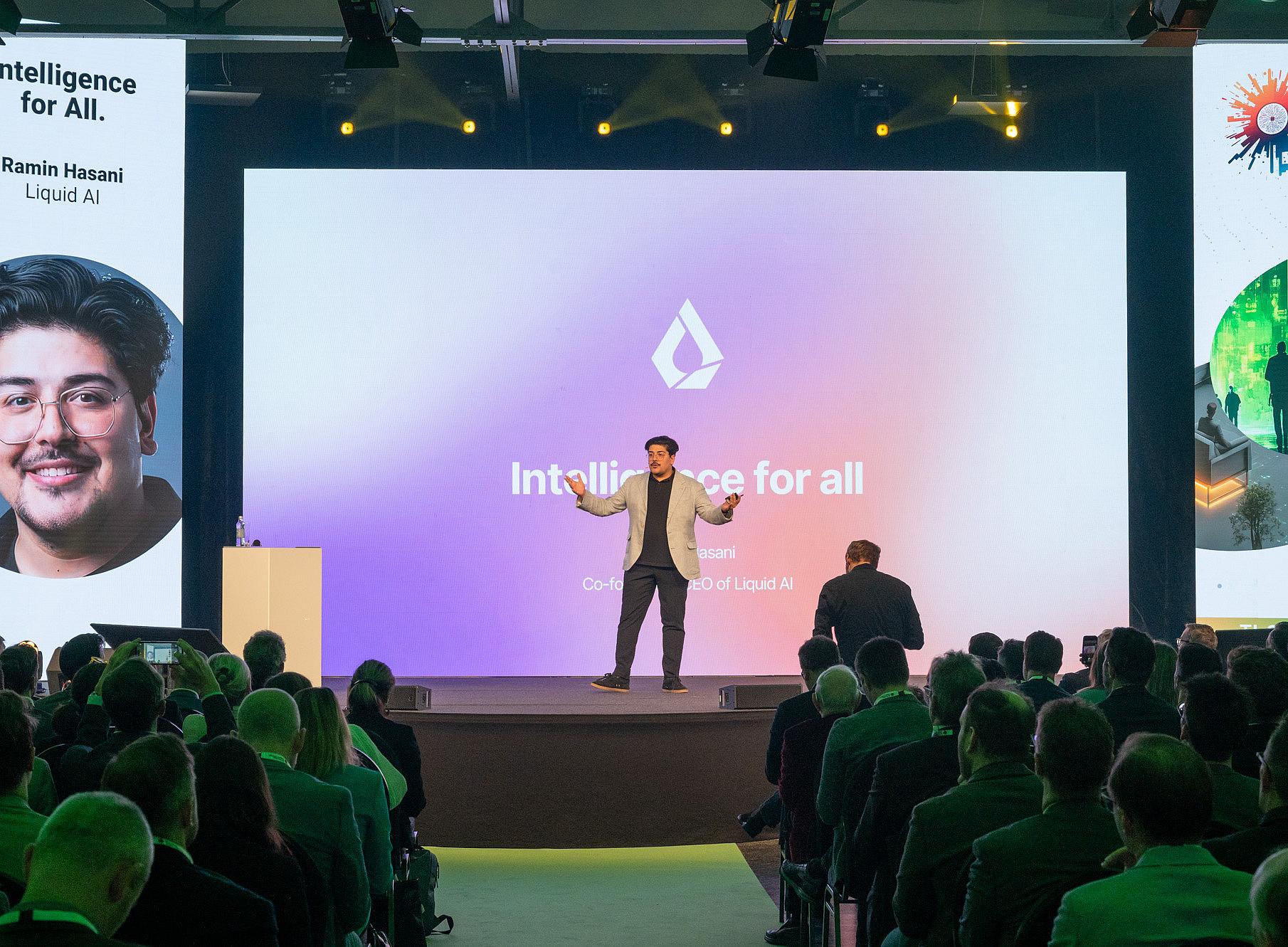 Ein Redner steht auf einer Bühne vor einem großen Bildschirm, der die Worte "Intelligence for all" und ein Logo zeigt. Der Hintergrund des Bildschirms ist in einem Farbverlauf von Lila zu Rosa gestaltet. Das Publikum sitzt in Reihen und hört dem Vortrag zu. Links und rechts der Bühne sind vertikale Banner mit Grafiken und Text angebracht. Die Beleuchtung der Bühne ist hell, während der Zuschauerbereich dunkler ist. 