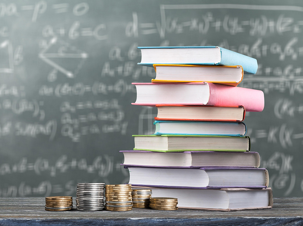 Thesis/Dissertation scholarship at the SBESS, A pile of books and five stacks of coins lie on a table and behind them is a blackboard with writing on it. ©BillionPhotos.com - stock.adobe.com