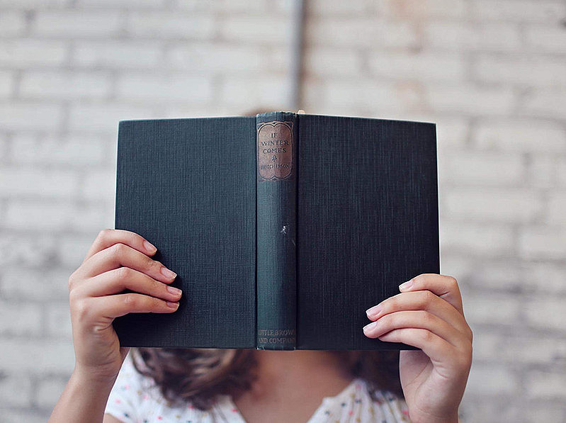 Eine Frau hält ein aufgeschlagenes Buch vor ihr Gesicht. 