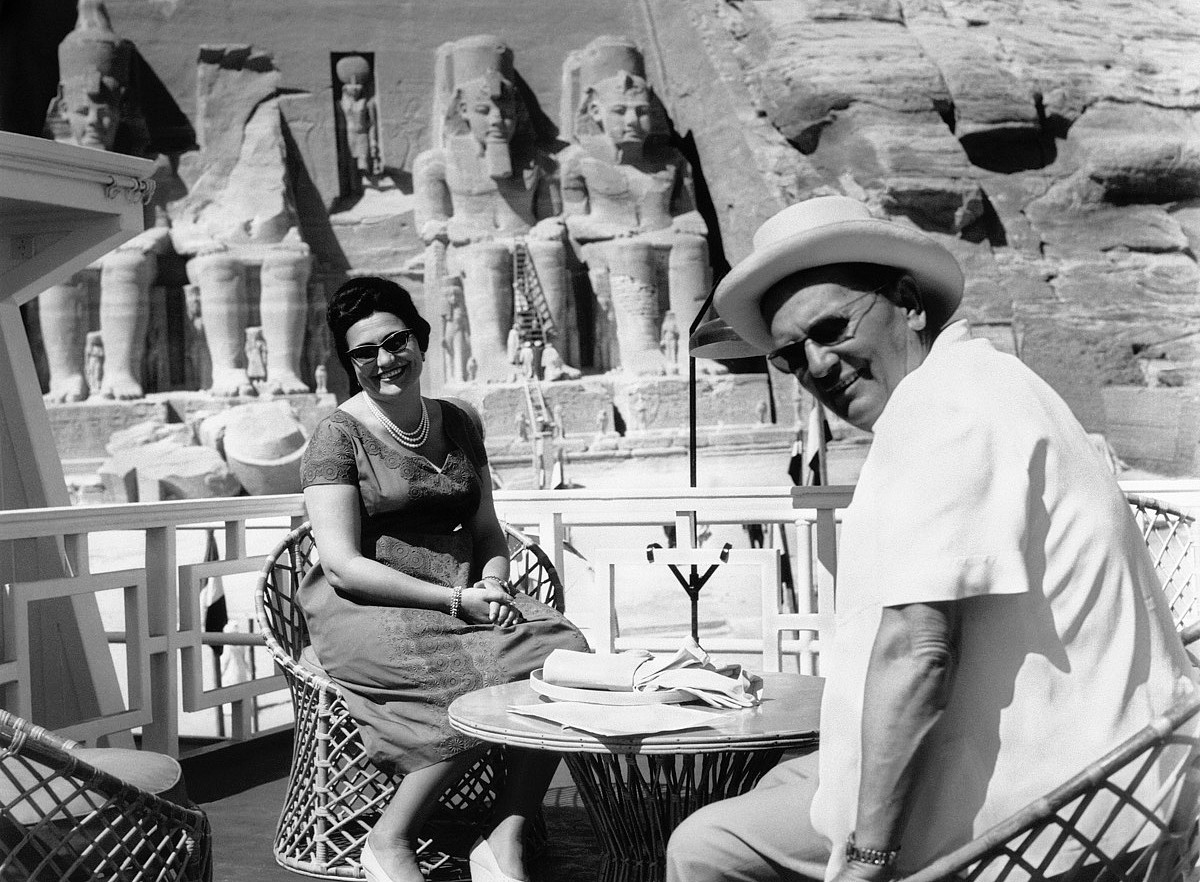 The black-and-white photograph shows two people sitting at a small round table on what appears to be the deck of a boat or a terrace. The woman is wearing a dress, jewelry, and heels, and is seated in a wicker chair. The man, dressed in light-colored clothing and a wide-brimmed hat, is sitting across from her. ©Fotoarchiv des Museums von Jugoslawien in Belgrad 