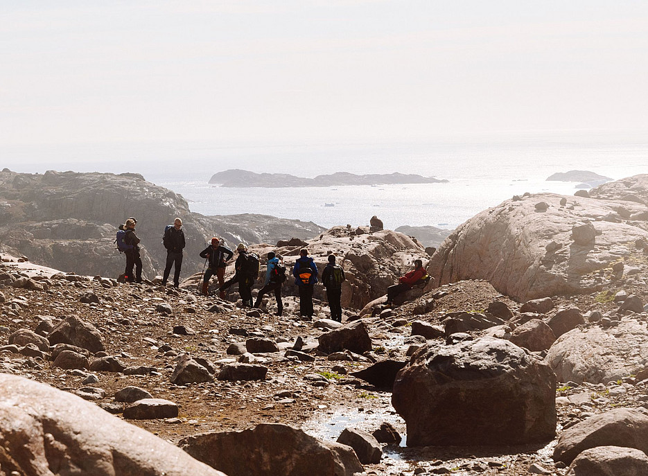 Eine Gruppe mit wandernden Menschen steht am felsigen Ufer Grönlands und blickt auf das Meer, umgeben von großen Felsen und kleinen Pfützen mit Regenwasser. Es ist ein sonniger Tag mit klarem Himmel. ©Karl Steinegger