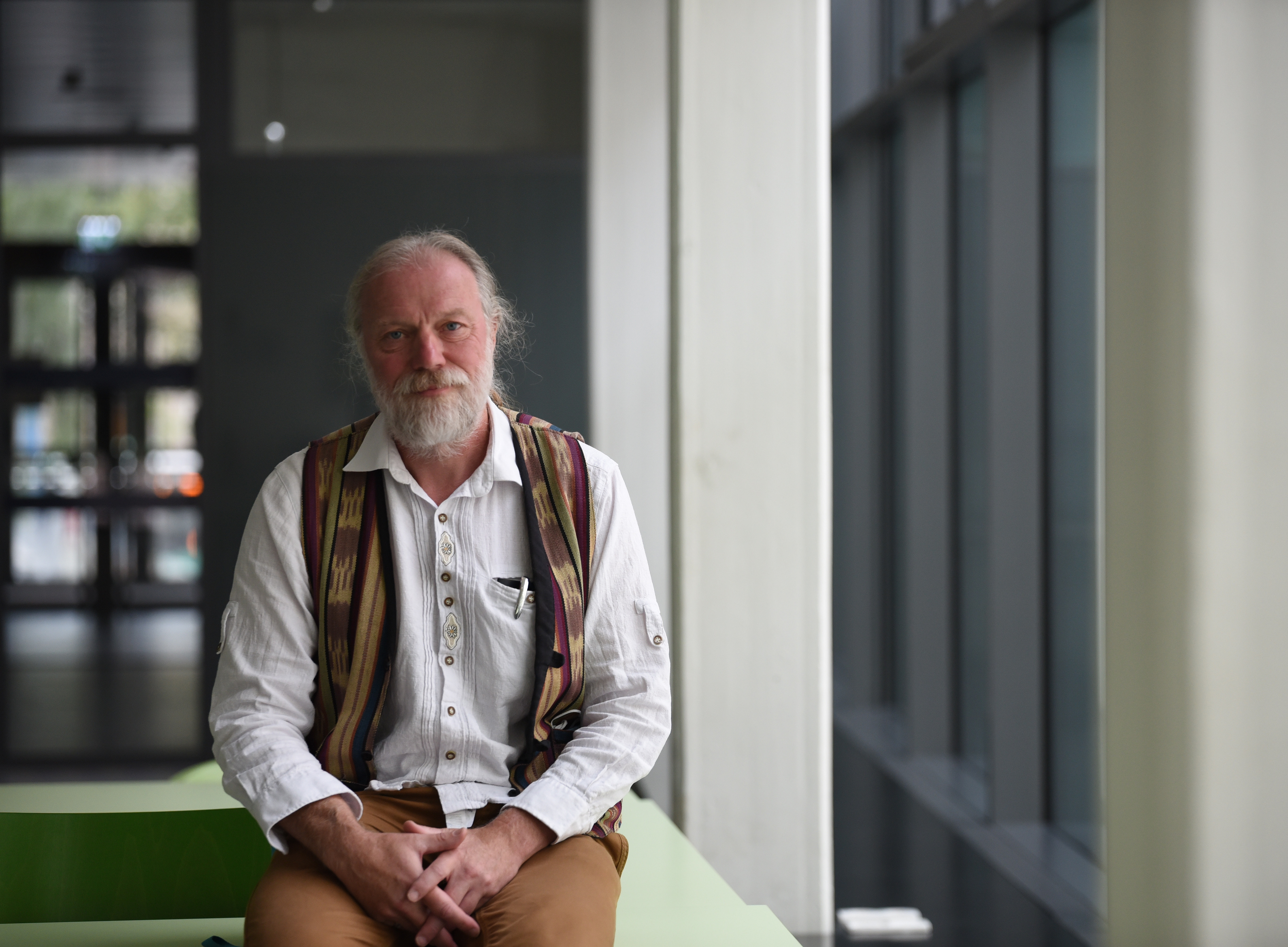 Ein Foto eines Professors mit grauem Haar und Bart, der an seiner Universität ein weißes Hemd und eine bunte Weste trägt und in einer Ganzkörperaufnahme in die Kamera lächelt 