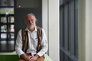 Ein Foto eines Professors mit grauem Haar und Bart, der an seiner Universität ein weißes Hemd und eine bunte Weste trägt und in einer Ganzkörperaufnahme in die Kamera lächelt