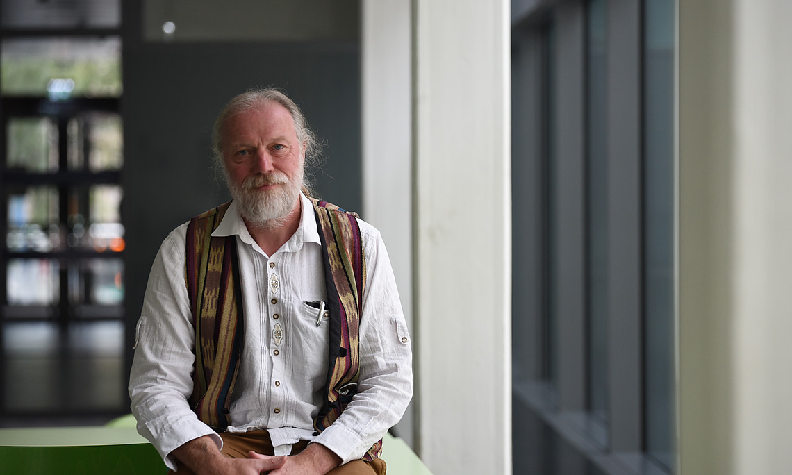 A photo of an professor with gray hair and beard, wearing a white shirt and colorful vest at his university, smiling for the camera in a full body shot