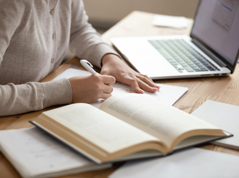 Bachelorarbeit an der SOWI-Fakultät, Person sitzt vor einem aufgeklappten Laptop und einem aufgeschlagenen Buch und arbeitet. ©fizkes - stock.adobe.com