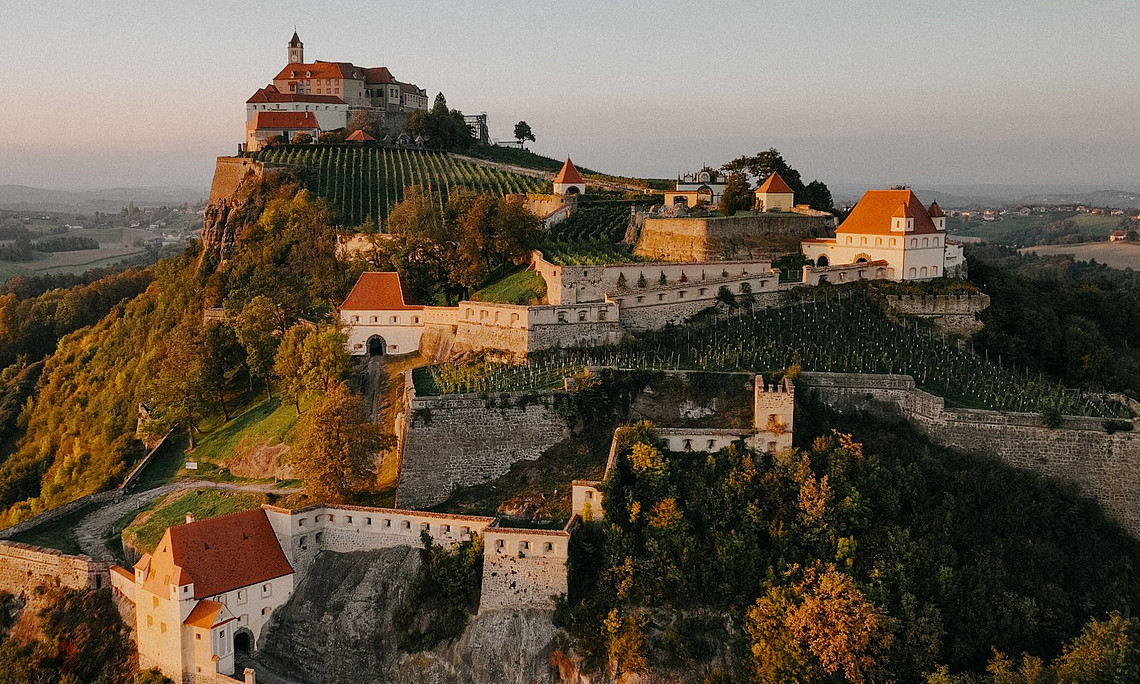 Eine historische Burganlage thront auf einem bewaldeten Hügel inmitten einer sanften Hügellandschaft. Die Burg besteht aus mehreren Gebäuden mit roten Dächern und ist von terrassenförmig angelegten Weinbergen und mächtigen Steinmauern umgeben. Der Sonnenuntergang taucht die Szene in warmes, goldenes Licht. Im Hintergrund erstreckt sich die ländliche Umgebung mit Feldern, kleinen Wäldern und vereinzelten Häusern bis zum Horizont.