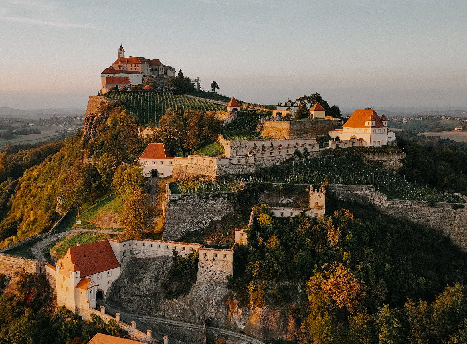 Eine historische Burganlage thront auf einem bewaldeten Hügel inmitten einer sanften Hügellandschaft. Die Burg besteht aus mehreren Gebäuden mit roten Dächern und ist von terrassenförmig angelegten Weinbergen und mächtigen Steinmauern umgeben. Der Sonnenuntergang taucht die Szene in warmes, goldenes Licht. Im Hintergrund erstreckt sich die ländliche Umgebung mit Feldern, kleinen Wäldern und vereinzelten Häusern bis zum Horizont. 