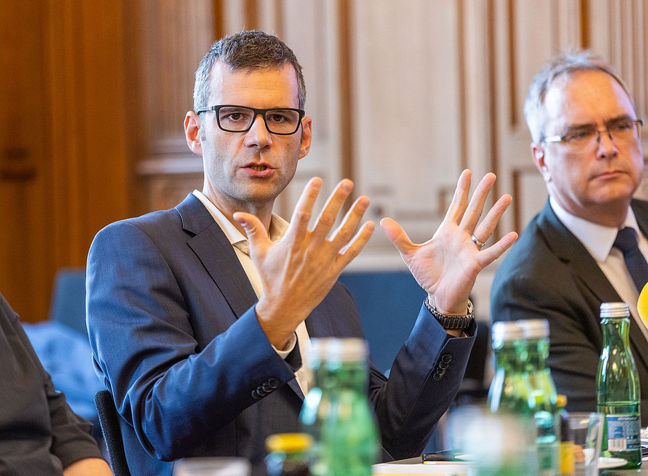 Fotografie eines Mannes mit Brille und Geschäftsanzug, der während einer Pressekonferenz über Finanzen an einem Tisch mit Kollegen spricht. Die Szene hat eine dramatische Wirkung, mit grünen Glasflaschen auf dem Tisch und den klaren Gesichtszügen der Person, die in einer ultra-realistischen Fotografie eingefangen wurden. ©www.photoworkers.at