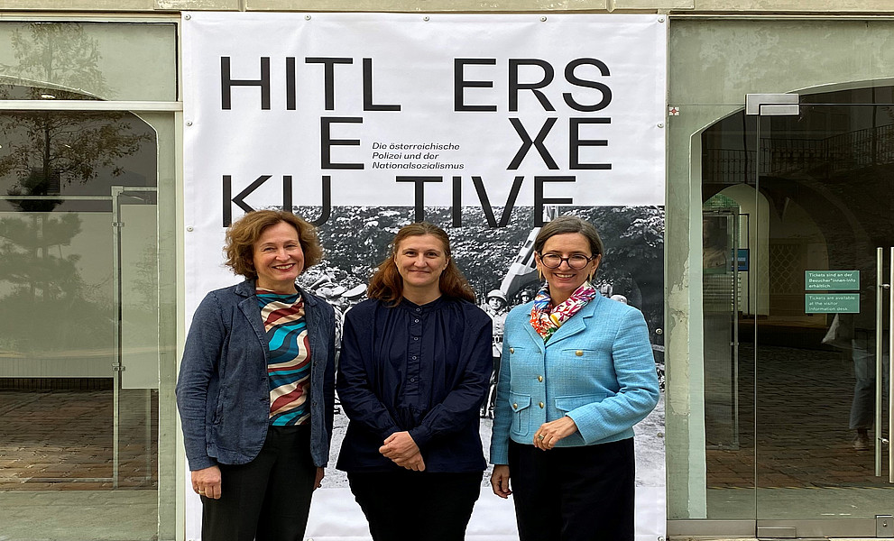 Drei Frauen stehen vor dem Eingang eines Kunstmuseums, lächeln und posieren für Fotos mit einem großen Banner im Rücken. Auf dem Banner steht „HITLERS Exekutive“.