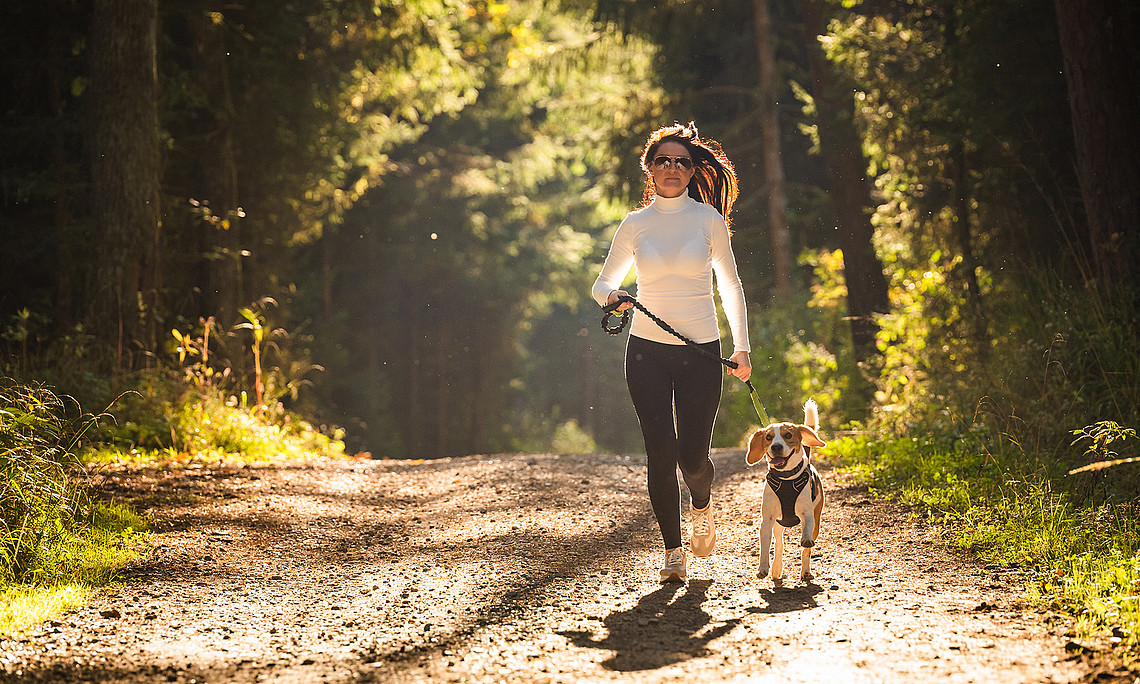eine Frau läuft mit einem Hund an der Leine durch den Wald