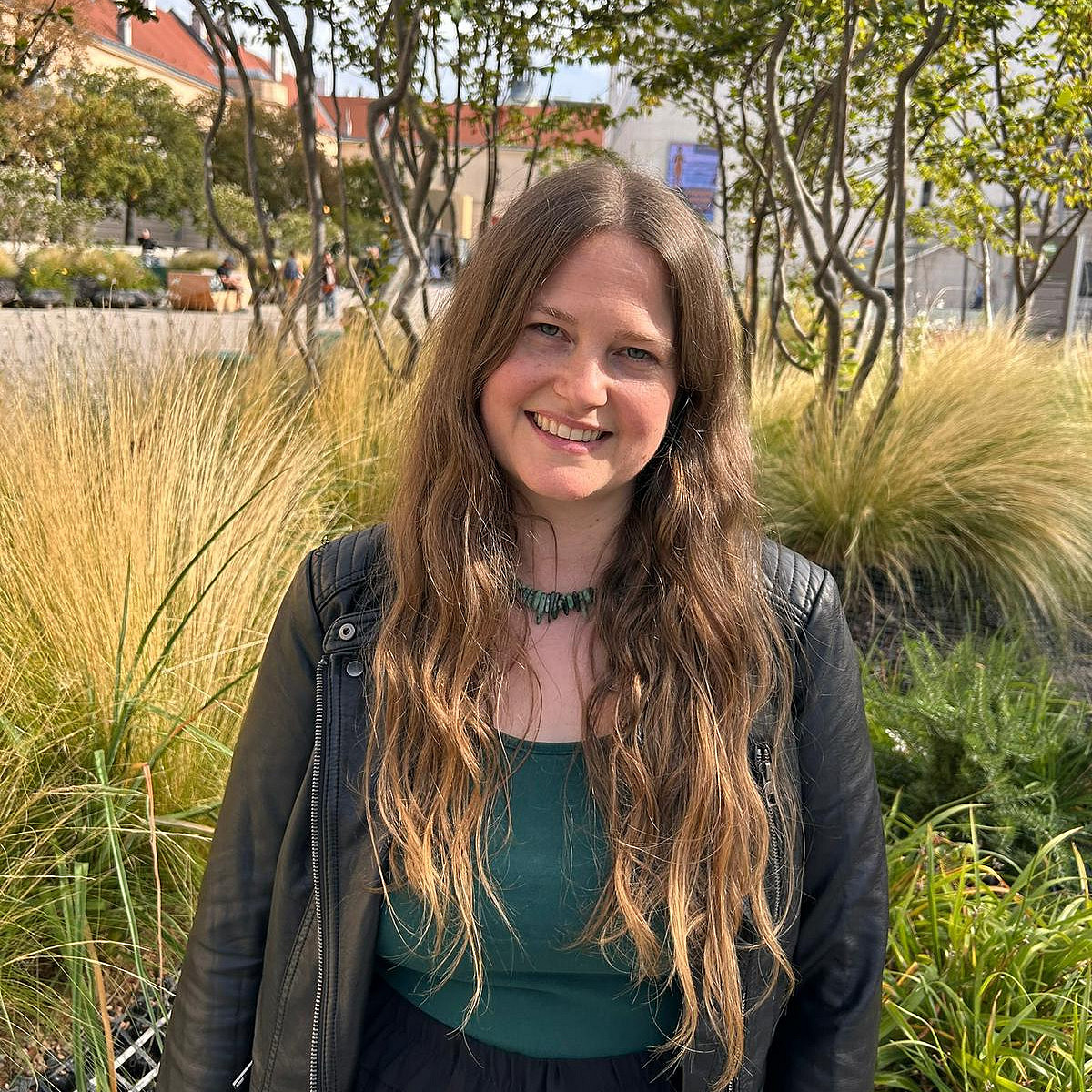 eine lächelnde junge Frau mit langen blonden Haaren und blauen Augen vor Büschen und Bäumen