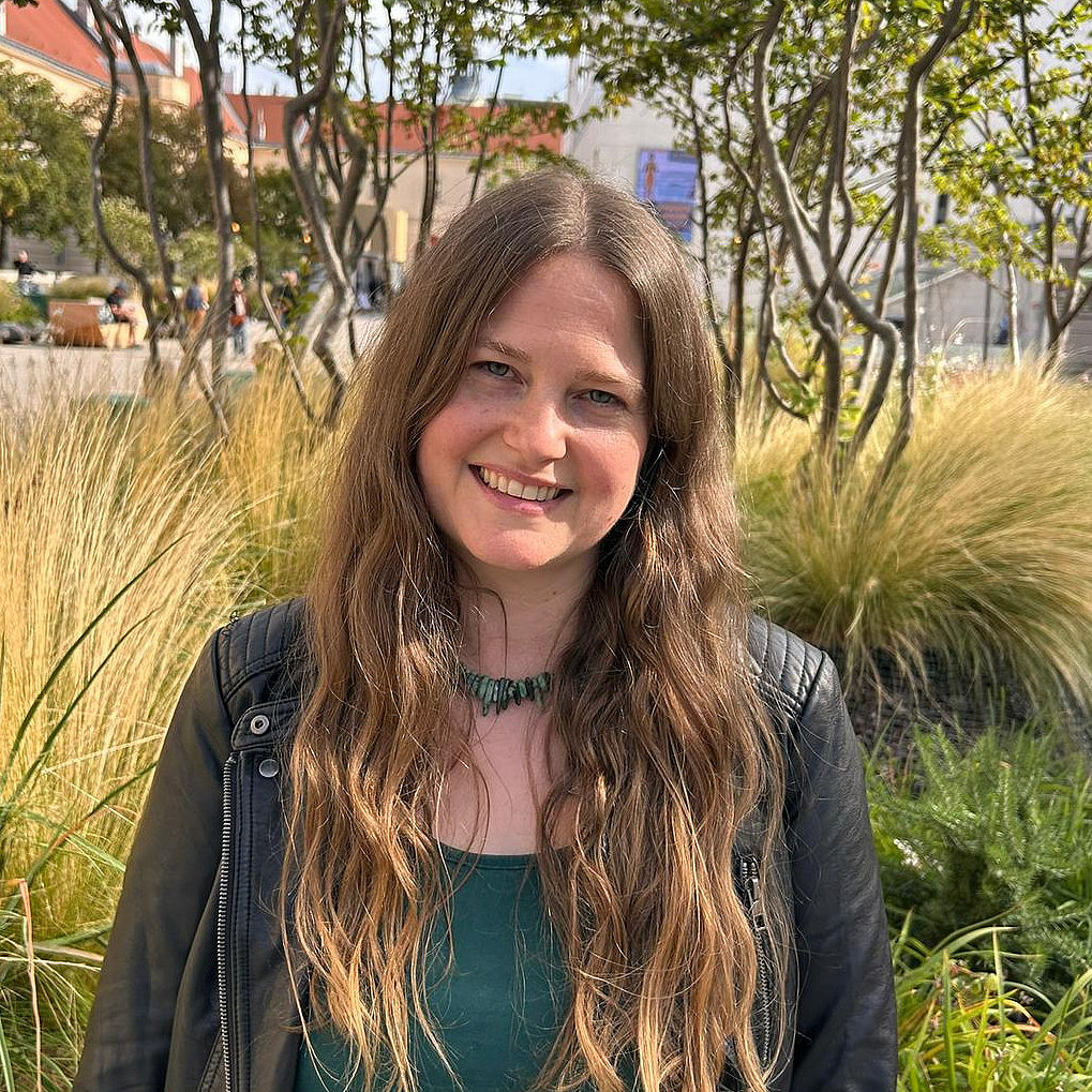 eine lächelnde junge Frau mit langen blonden Haaren und blauen Augen vor Büschen und Bäumen