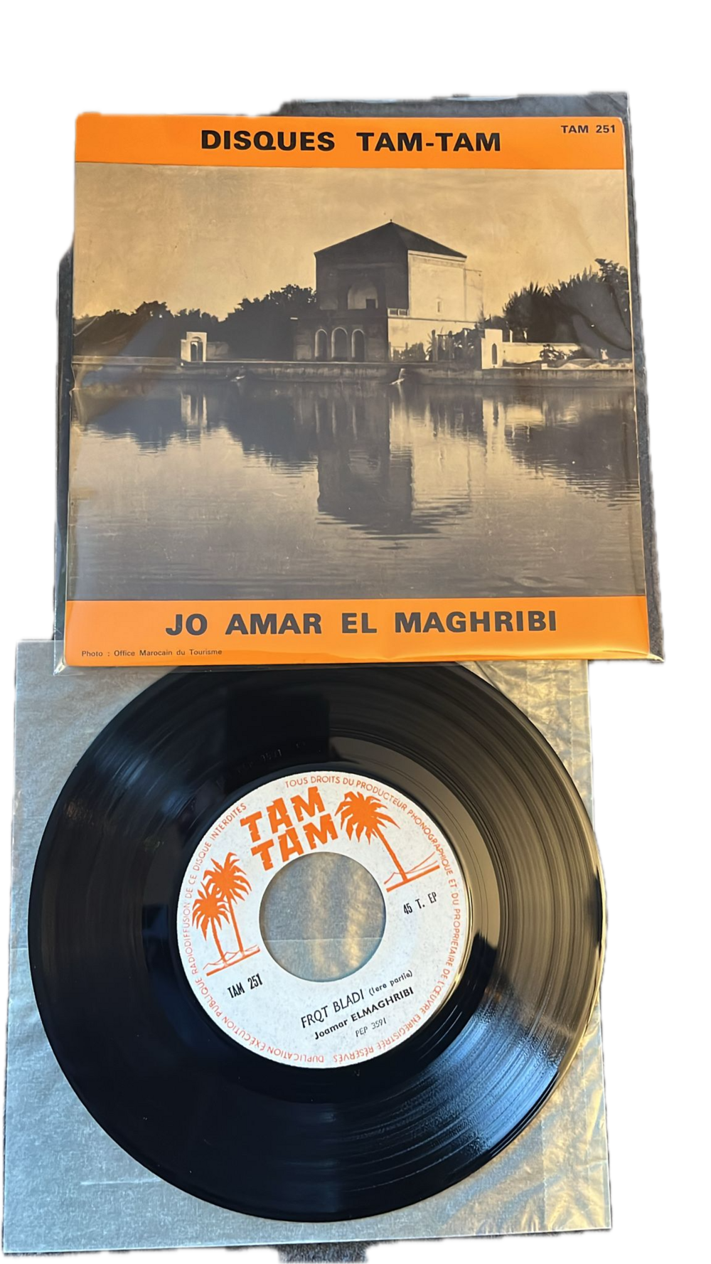 Das Foto zeigt eine schwarze Vinylplatte auf einem hellen Untergrund, deren Cover eine Schwarz-Weiß-Fotografie eines Gebäudes am Wasser zeigt. Auf dem Cover steht in orangefarbener und weißer Schrift „DISQUES TAM-TAM” und „JO AMAR EL MAGHRIBI”. ©Gharamophone