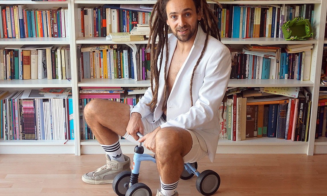 Autor Max Höfler auf einem Kleinkinder-Fahrzeug vor einem Bücherregalvor
