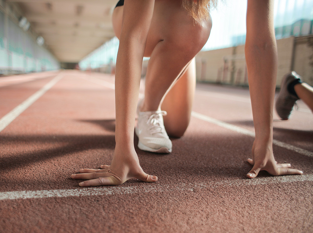 Zu sehen sind die Beine und Arme einer Frau in weißen Turnschuhen und kurzer Hose an der Startlinie auf einer Laufbahn. Die Person ist in Startposition. ©pexels_andrea piacquadio
