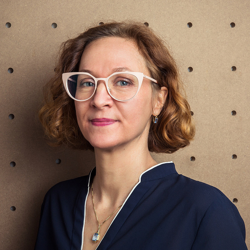 Das Foto von Astrid Kury, einer Frau mit lockigem Haar, die eine große weiße Brille und eine marineblaue Bluse trägt und vor einem strukturierten braunen Hintergrund mit Perforationen posiert. ©Karin Lernbeiß