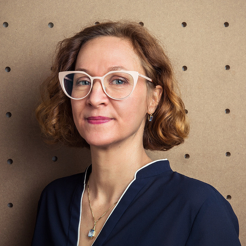 Das Bild zeigt ein Porträt von Astrid Kury, einer Frau mit lockigem, rotbraunem Haar und blauen Augen. Sie trägt eine weiße Brille mit katzenaugenförmigem Rahmen und ein marineblaues Oberteil mit weißem Kragen. Sie trägt auch eine goldene Halskette mit einem blauen Anhänger und passende Ohrringe. Der Hintergrund ist eine hellbraune Wand mit einem regelmäßigen Muster aus dunklen Löchern. Die Beleuchtung ist gleichmäßig und hell, was die Details von Gesicht und Kleidung hervorhebt. ©Karin Lernbeiß