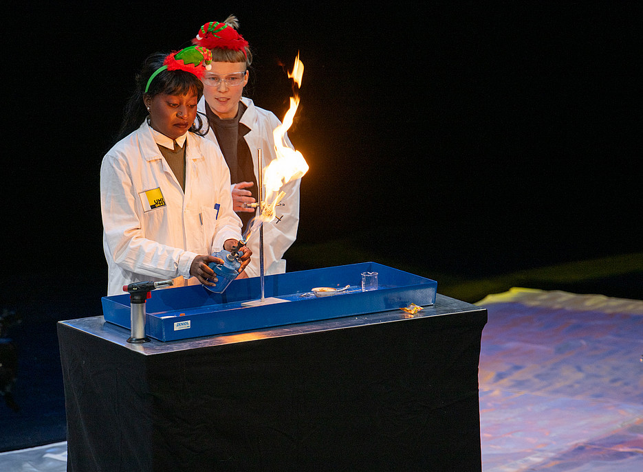 Zwei Personen in weißen Laborkitteln stehen hinter einem Tisch mit schwarzer Abdeckung, auf dem ein blaues Tablett mit Laborutensilien wie einem Becherglas und einem Löffel liegt; eine Person hält einen Gasbrenner, aus dem eine Flamme aufsteigt. 