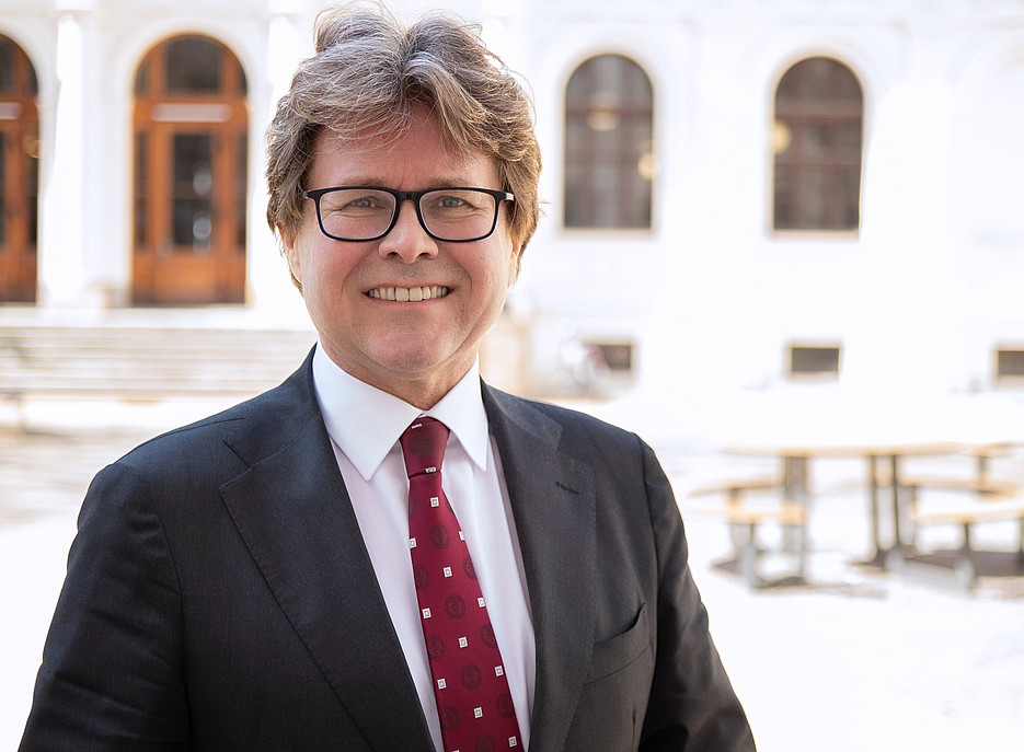 Ein Mann in einem dunklen Anzug mit weißem Hemd und roter Krawatte steht im Freien vor einem historischen Gebäude mit weißen Fassaden und großen, bogenförmigen Fenstern. Im Hintergrund sind Holztische und Bänke auf einem gepflasterten Platz zu sehen. 