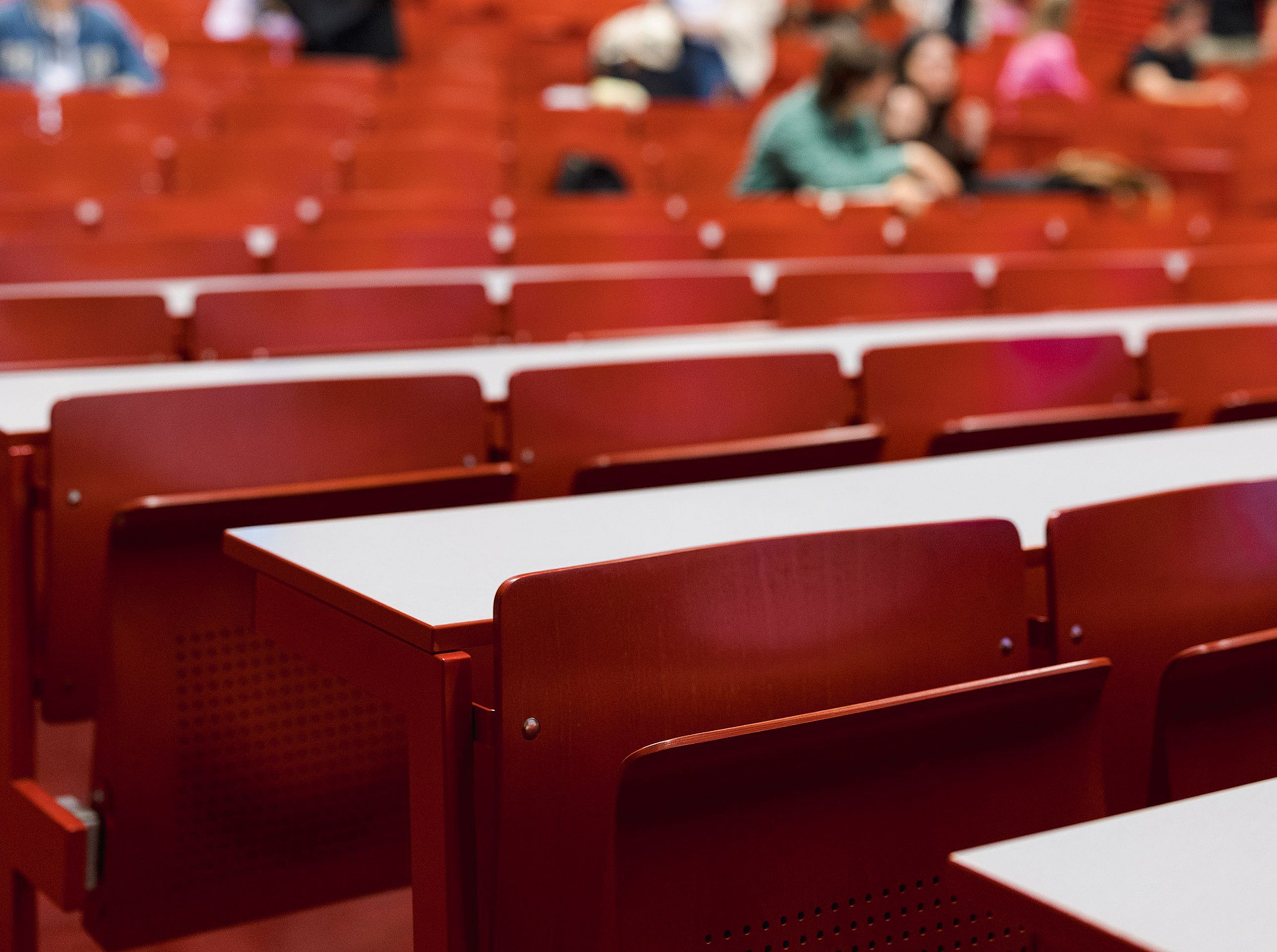 Auf dem Bild ist ein Hörsaal mit roten Stühlen zu sehen, während die Studierenden im Hintergrund verschwommen dargestellt sind. ©Uni Graz/Kanizaj