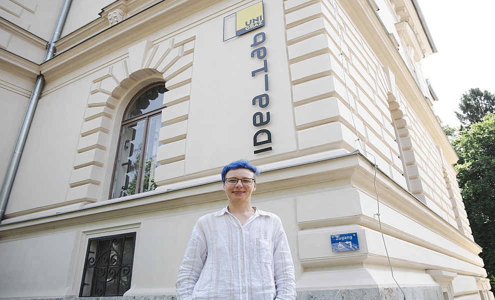 Eine junge Frau mit kurzen blauen Haaren steht vor einem alten Gebäude und lächelt in die Kamera. Auf dem Gebäude steht „IdeaLab“. Sie trägt ein weißes Leinenhemd, Jeans und eine Brille. 