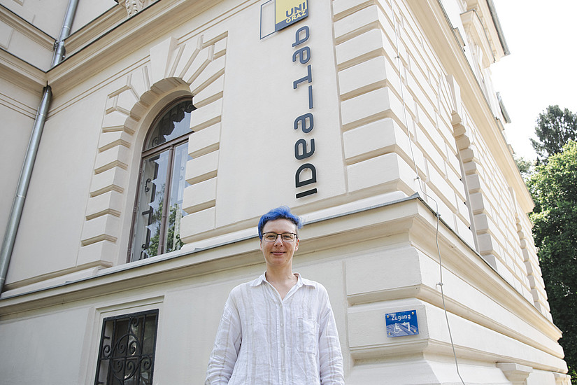 Eine junge Frau mit kurzen blauen Haaren steht vor einem alten Gebäude und lächelt in die Kamera. Auf dem Gebäude steht „IdeaLab“. Sie trägt ein weißes Leinenhemd, Jeans und eine Brille. 