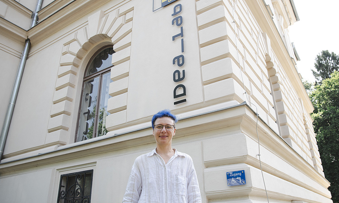 Eine junge Frau mit kurzen blauen Haaren steht vor einem alten Gebäude und lächelt in die Kamera. Auf dem Gebäude steht „IdeaLab“. Sie trägt ein weißes Leinenhemd, Jeans und eine Brille. 