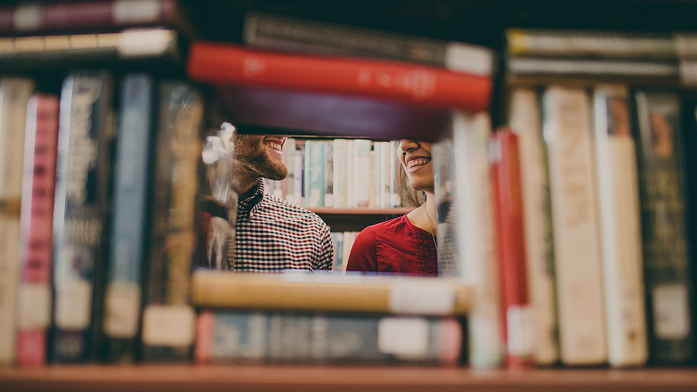 Bücher in einem Regal bilden ein kleines Fenster, durch dass man zwei Personen sieht, die sich einander zuwenden und lächeln. ©(c) Josh Felise auf Unsplash