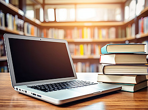 Ein aufgeklappter Laptop und ein Bücherstapel liegen auf einem Tisch in einer Bibliothek ©Marat - stock.adobe.com