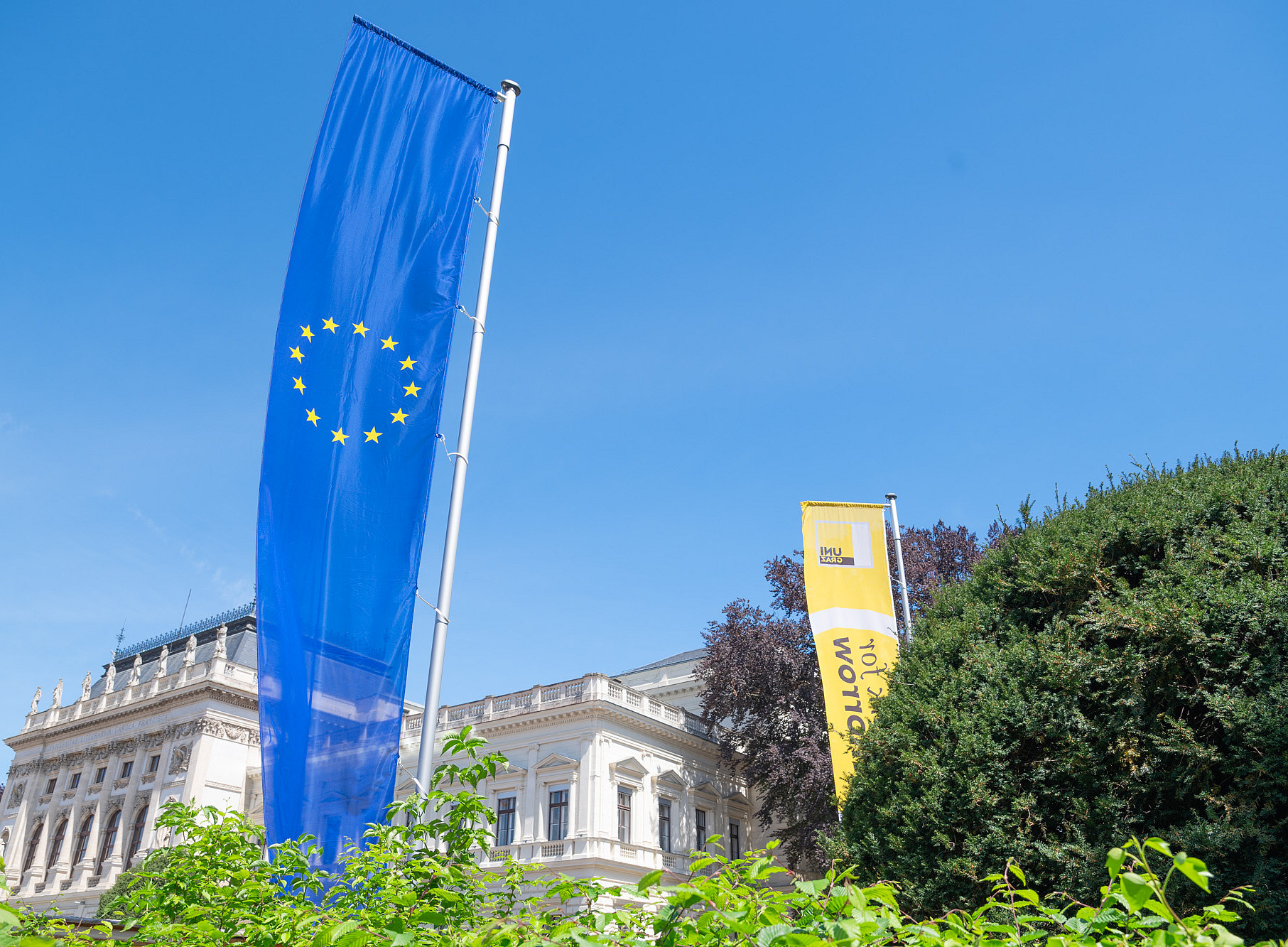 EU-Fahne und gelbe Fahne mit Uni Graz-Logo vor historischem Gebäude und Grünanlage ©Uni Graz/Angele