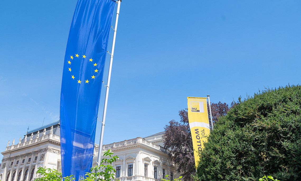EU-Fahne und gelbe Fahne mit Uni Graz-Logo vor historischem Gebäude und Grünanlage