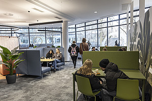 A modern lounge with large windows that let in plenty of daylight. Several people are sitting on green and blue seating areas, some chatting, others playing board games. In the foreground is a green plant in a black pot. The wall on the right is decorated with large plant motifs. In the background, you can see more seating areas and a bright, open atmosphere.