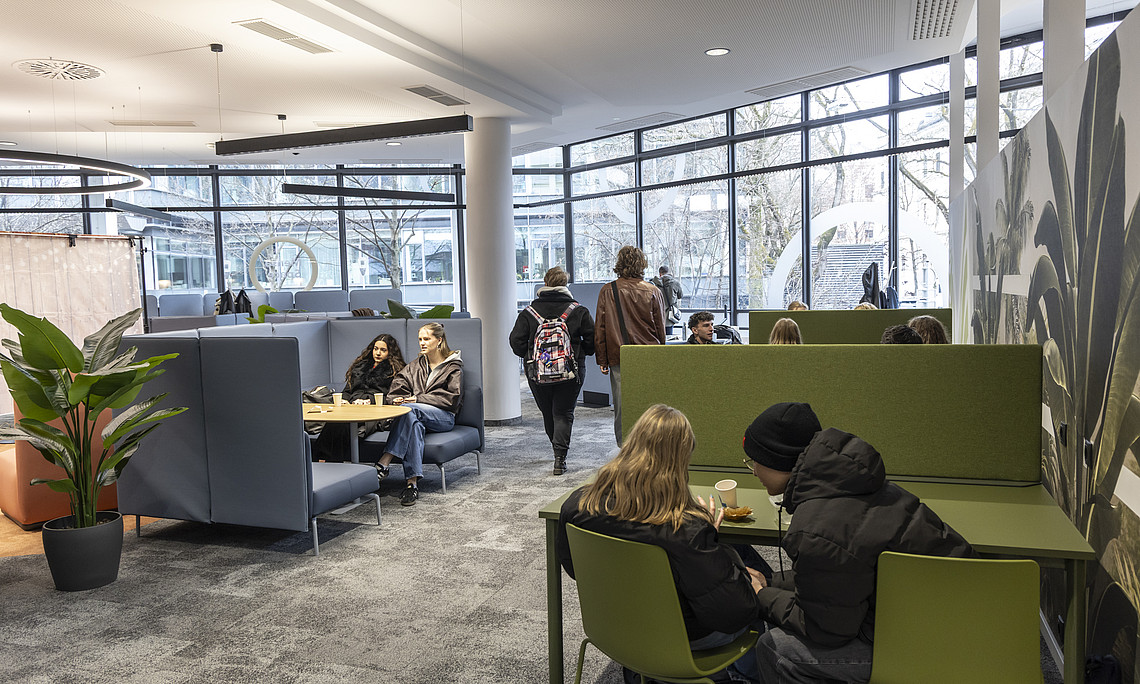A modern lounge with large windows that let in plenty of daylight. Several people are sitting on green and blue seating areas, some chatting, others playing board games. In the foreground is a green plant in a black pot. The wall on the right is decorated with large plant motifs. In the background, you can see more seating areas and a bright, open atmosphere.