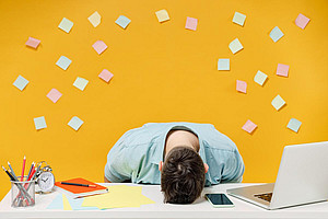 A person's head is slumped on their desk. The head is lying on the surface of the desk, with a laptop, notepad and writing materials next to it. Behind the person is a yellow wall full of Post-It notes. ©ViDi Studio - stock.adobe.com