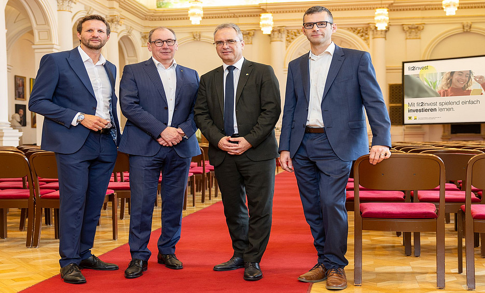 Ein Foto von vier Männern in Anzügen, zwei mit und einer ohne Brille, die auf dem roten Teppich in der Aula der Universität Graz stehen, im Hintergrund ist ein Bildschirm zu sehen, der Produkte des Unternehmens zeigt. Davor steht eine Stuhlreihe mit drei Stühlen nebeneinander, 