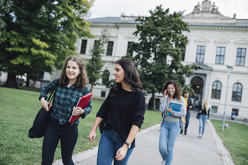 2 Studierende spazieren über den Campus Richtung Hauptgebäude und unterhalten sich miteinander; im Hintergrund sind weitere Studierende zu sehen; neben einem alten schönen Gebäude der Uni Graz ist eine Wiese mit Bäumen in sattem Grün zu sehen