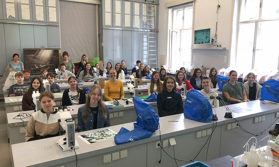A group of students in a German high school laboratory, sitting at their desks and smiling for a photo with blue-bagged science supplies on top of each desk. In front is an electric microscope that shines light into a small plastic box filled with organic material. 